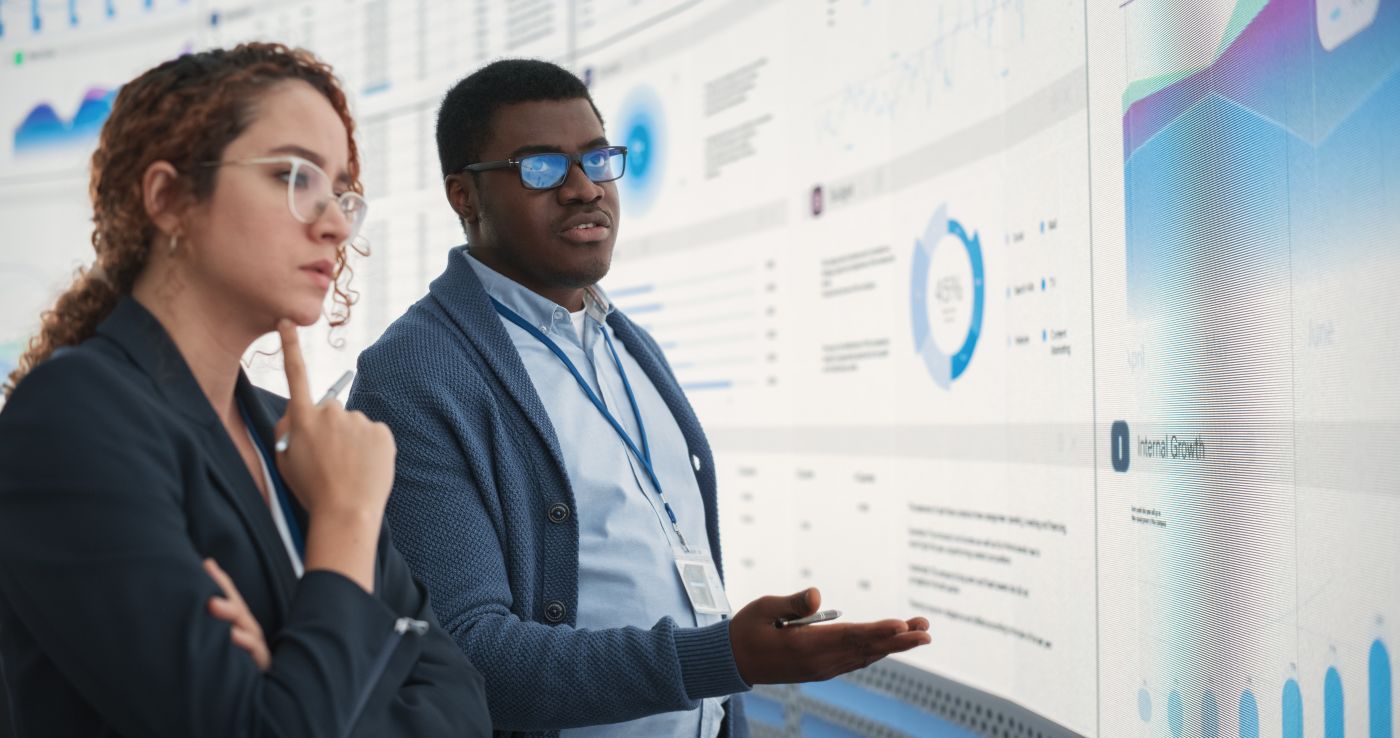 Black Man And Hispanic Woman Working On Business Strategy And Standing Infront Of Big Digital Screen With Data. Male And Female Colleagues Discussing Plan For Profit Margins Enlargement In The Office. - Auster Inteligência Contábil