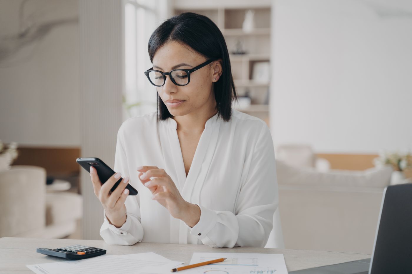 Businesswoman In Glasses Using Mobile Phone Apps For Business Time Management Sitting At Laptop - Auster Inteligência Contábil