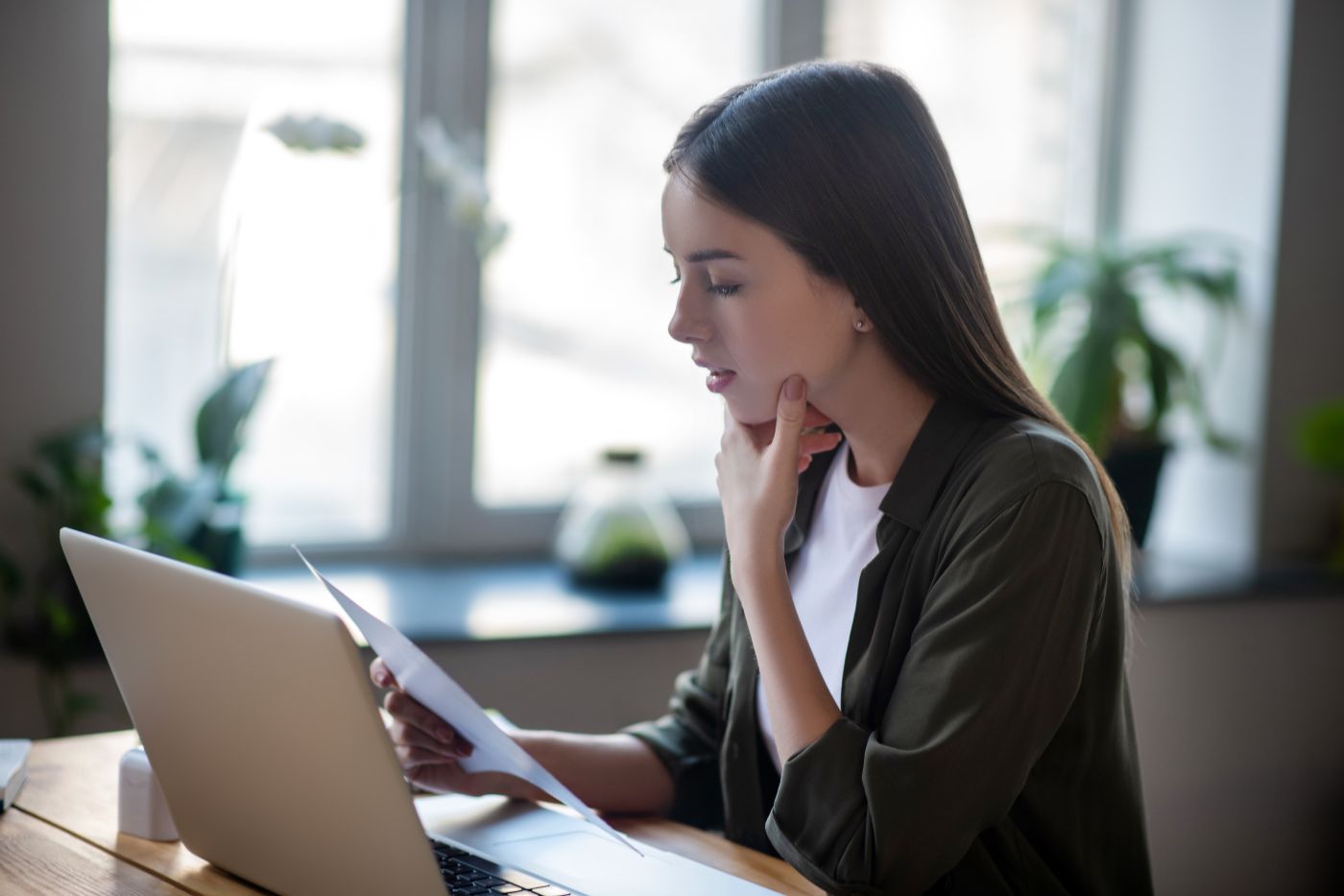 Girl Sitting In Front Of Laptop Carefully Looking At Paper Document. - Auster Inteligência Contábil